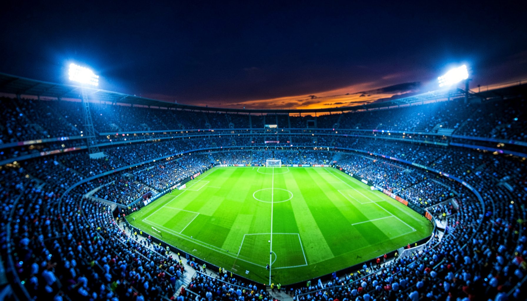 Wide aerial view of a packed stadium at dusk, floodlights blazing across a vibrant green pitch, deep navy sky with amber and electric-blue reflections